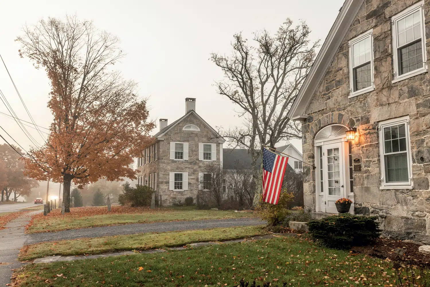 Stone house with an American flag by the front door, trees with autumn leaves, and cloudy sky. Nearby road and another stone building in the background.