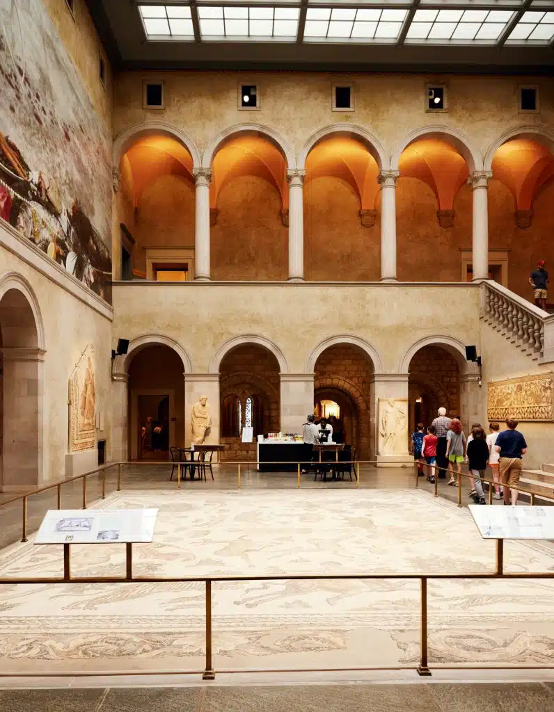 A museum interior with arches, columns, and a large mural. People are walking and observing exhibits, including a mosaic on the floor.