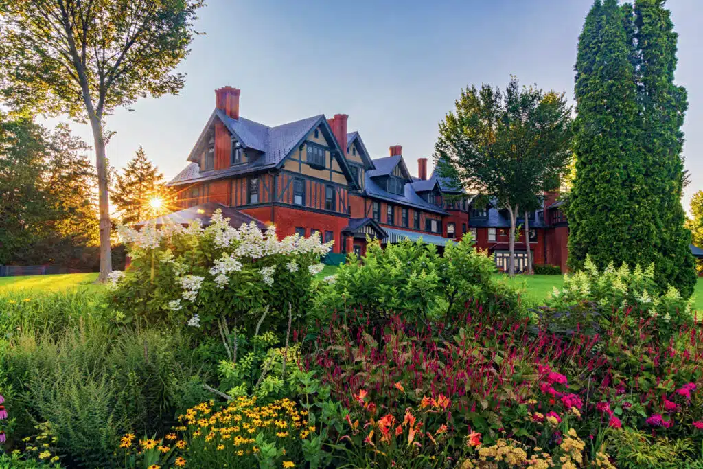 A large, red brick and timber house stands behind a colorful flower garden and green shrubs at sunset, with tall trees in the background.