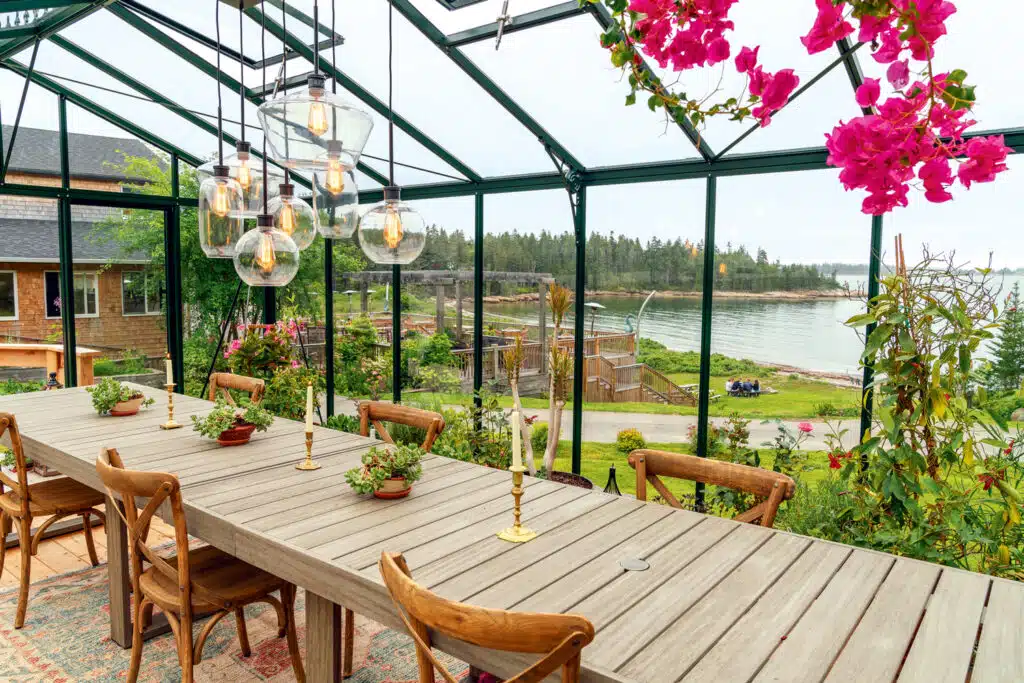 A long wooden table with chairs sits inside a glass greenhouse with hanging lights, overlooking a scenic view of water, trees, and a house. Pink flowers hang from above.