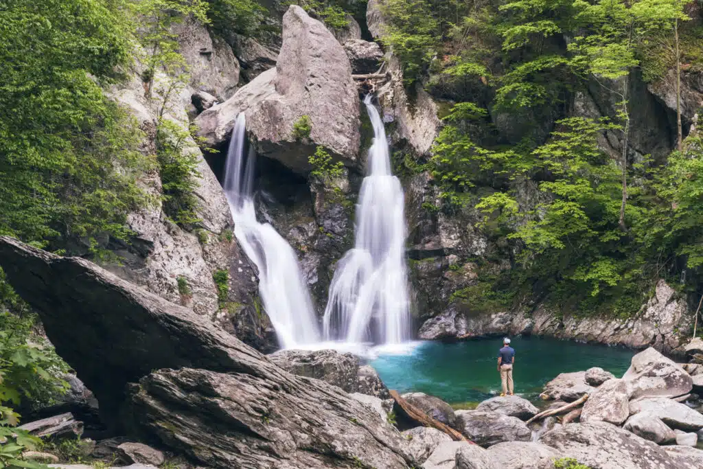 A person stands on rocks near a small waterfall flowing into a clear pool, surrounded by dense green forest and large boulders.