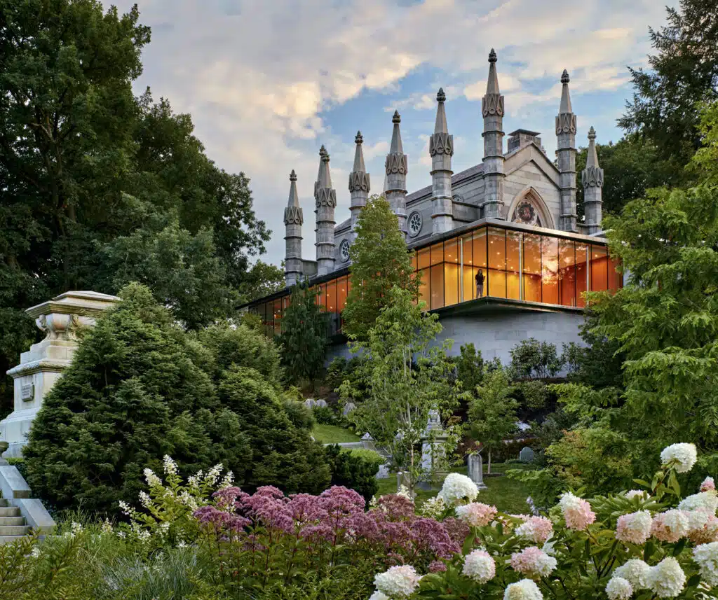 A modern glass-walled building adjoins a historic Gothic-style structure, surrounded by lush gardens and flowering shrubs under a partly cloudy sky.