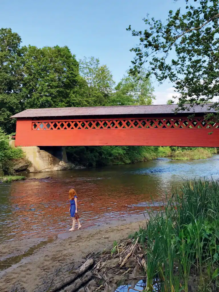 A child stands at the edge of a shallow creek near a red covered bridge surrounded by green trees and foliage.