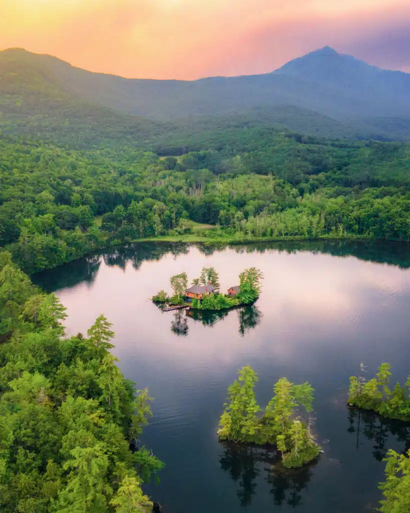 A small house sits on a tree-covered island in the middle of a calm lake, surrounded by dense forest and mountain ranges under a colorful sky.