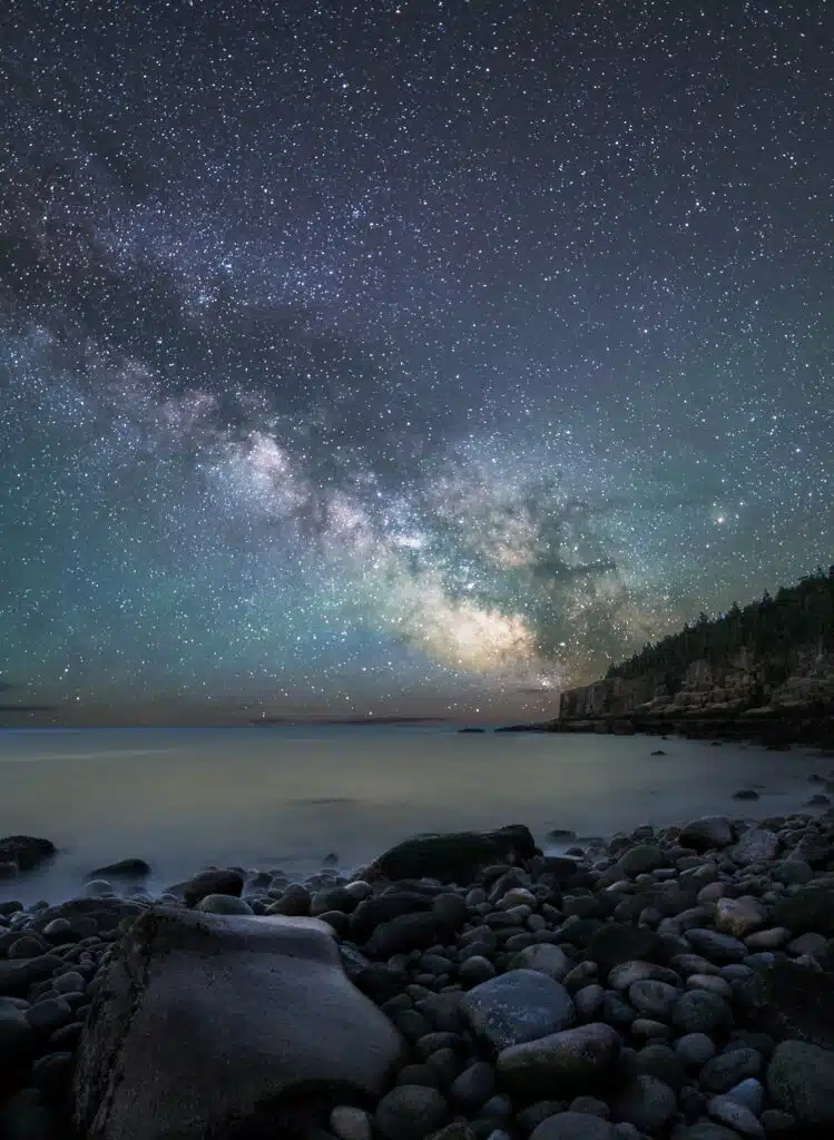 Rocky coastline at night with the Milky Way galaxy visible in a star-filled sky above, cliffs and forest in the background.