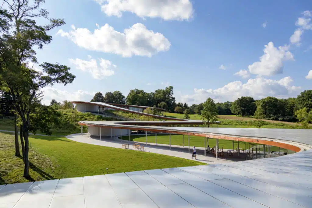 Modern, curved glass building with a green roof set in a grassy landscape with trees, outdoor seating, and a blue sky with scattered clouds.