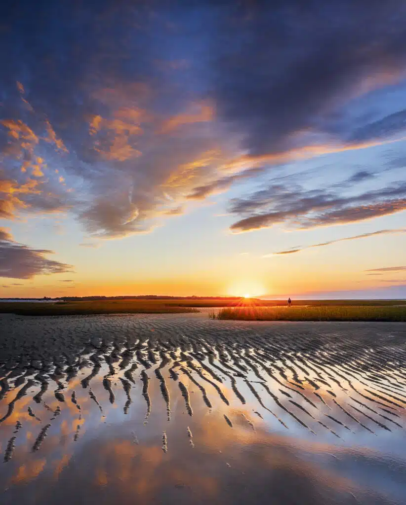 A sunset over a sandy beach with rippled wet sand reflecting clouds in the sky; a person stands in the distance near grassy dunes.