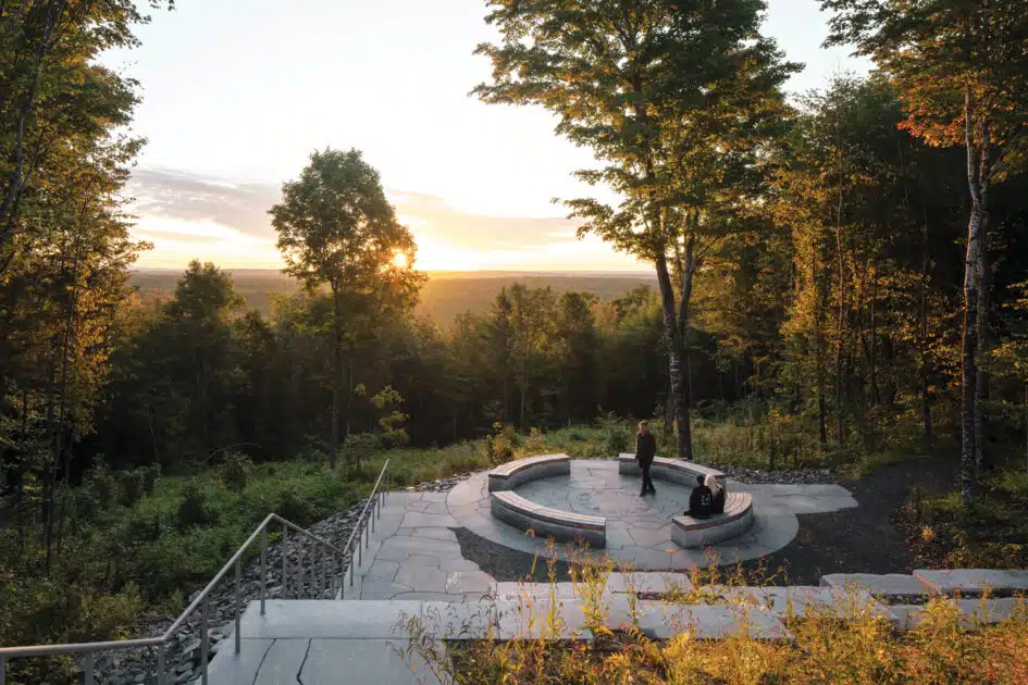 A circular stone seating area overlooks a forested landscape at sunset, with two people sitting on the benches surrounded by tall trees. 2025 Maine Travel Guide | Hotels, Dining & Attractions