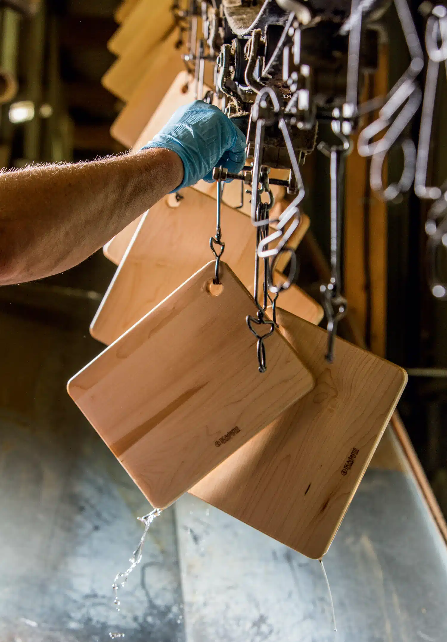 A hand in a blue glove holds a wooden board with hooks in a drying line with several similar boards hanging. Water droplets are visible on the surface.